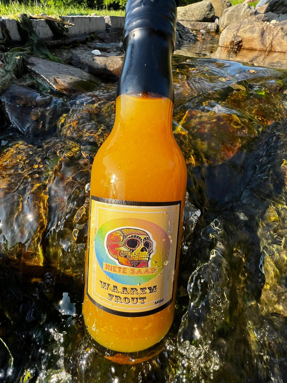Bouteille de sauce piquante Waarem Frout posée sur des rochers avec eau courante, marquée par des saveurs d'ananas et de mangue épicée.